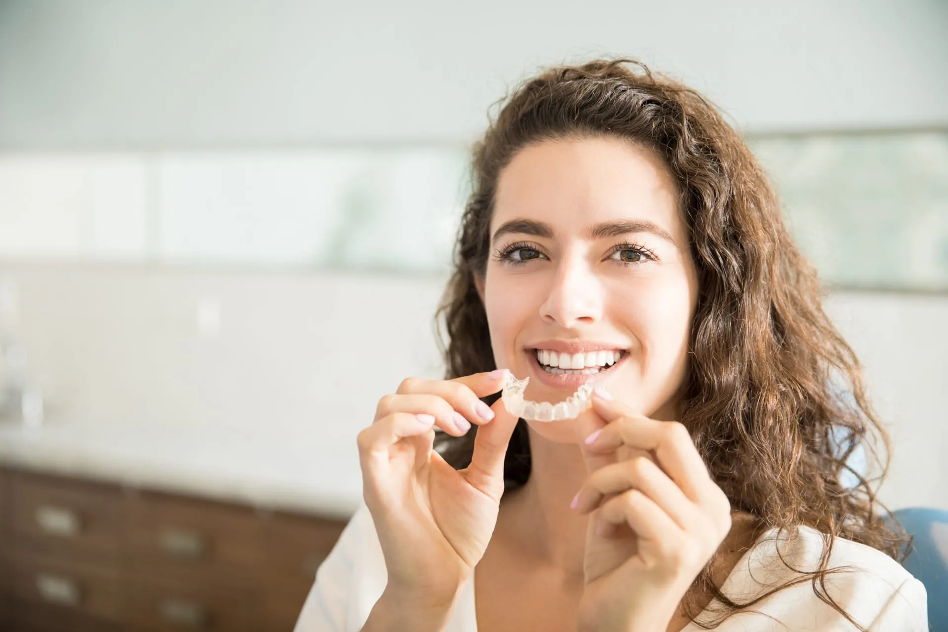 A brunette woman with a warm smile holding a clear retainer in her hands, showcasing an orthodontic accessory for teeth alignment.